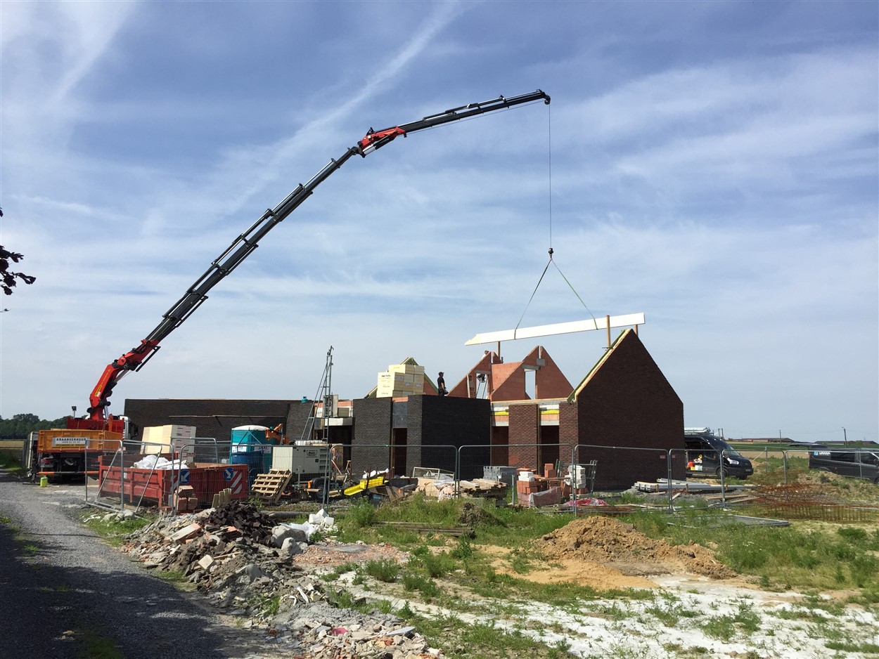 Kraanwagen met uitgeschoven giek hijst een dakbalk boven een half afgewerkte rijwoning in aanbouw